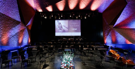 Sala koncertowa z ustawionymi krzesłami i pulpitami dla orkiestry, z przodu stoi bukiet kwiatów w koszyku a z tyłu na ekranie wyświetla się nazwa pierwszego etapu konkursu