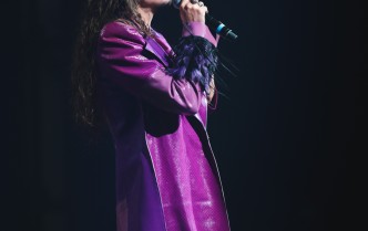 mężczyzna w fioletowym płaszczu śpiewający do mikrofonu, wokół czarne tło