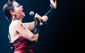 kobieta w czarno-czerwonej sukni śpiewająca do mikrofonu z wyciągniętą w górę lewą ręką