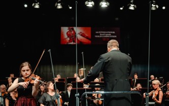 kobiera grająca na skrzypcach, obok stoi dyrygent, w tle siedzi orkiestra